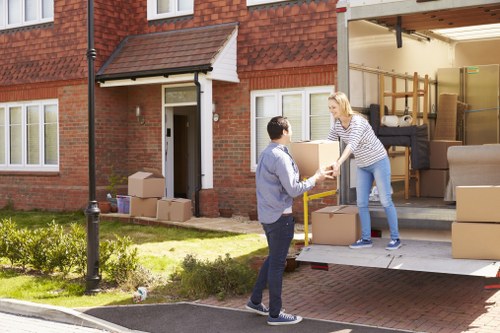A woman with blonde hair wearing a striped long-sleeve top and light blue jeans is standing inside a moving van parked outside a brick residential house in Lambeth. She is handing a cardboard box to a man with short dark hair, dressed in a grey jacket and dark trousers, who is standing on the driveway. The open rear doors of the van reveal several packed cardboard boxes of various sizes and some furniture, including a rolled-up carpet and a wooden shelving unit. The house’s exterior features a red-brick facade with white window frames and a small canopy above the front door. A grassy garden with small plants and a lamppost is visible on the left side of the driveway. The scene is set during daytime with natural light illuminating the environment, capturing the process of a local house relocation carried out by Lambeth Man with Van, providing professional removals services in the area.